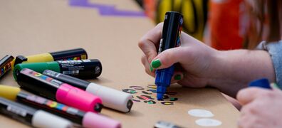 Hand mit grüner Nagellackierung hält einen blauen Marker und malt auf braunes Papier, daneben liegen mehrere bunte Marker.