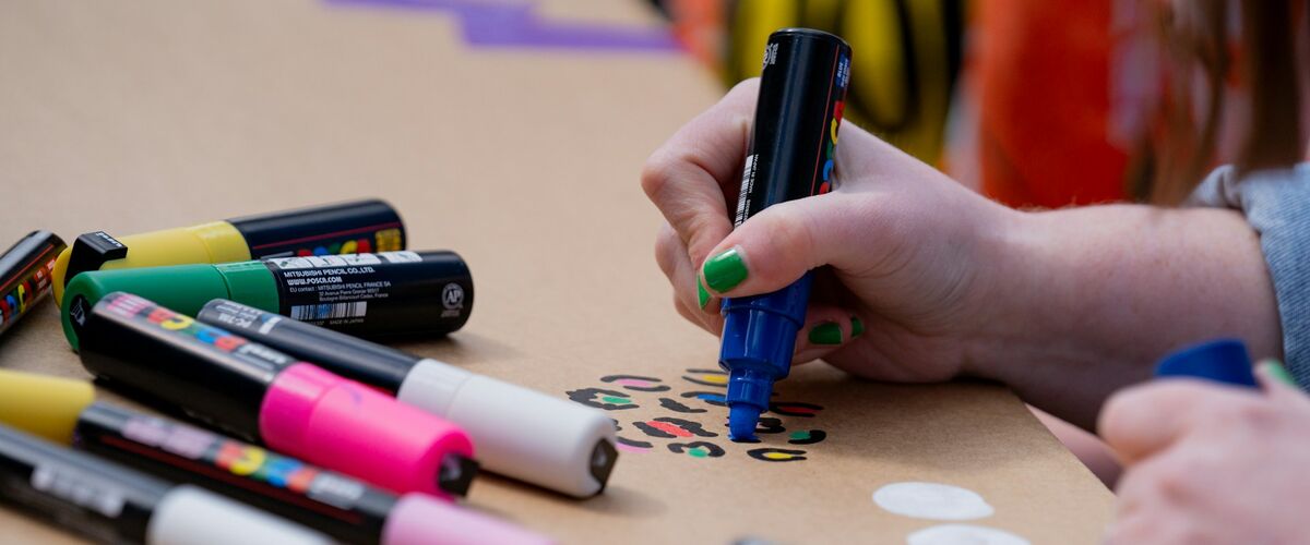 Hand mit grüner Nagellackierung hält einen blauen Marker und malt auf braunes Papier, daneben liegen mehrere bunte Marker.