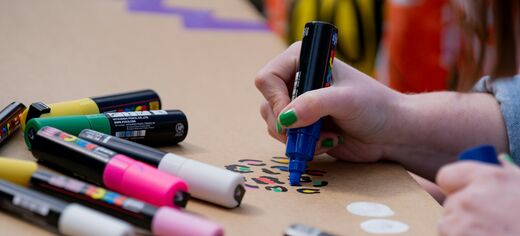 Hand mit grüner Nagellackierung hält einen blauen Marker und malt auf braunes Papier, daneben liegen mehrere bunte Marker.