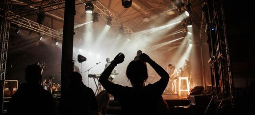 Symbolbild: Silhouette von Konzertbesuchern vor einer beleuchteten Bühne mit Musikern und Lichtstrahlen
