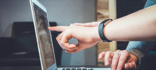 Hand zeigt auf Laptopbildschirm, andere Hand tippt auf Tastatur