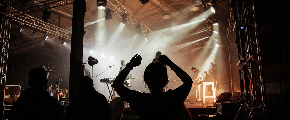 Symbolbild: Silhouette von Konzertbesuchern vor einer beleuchteten Bühne mit Musikern und Lichtstrahlen