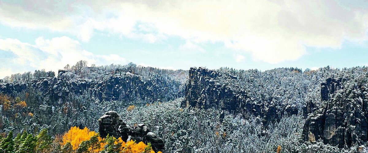 Winterlandschaft mit schneebedeckten Bergen und bunten, herbstlichen Bäumen im Vordergrund